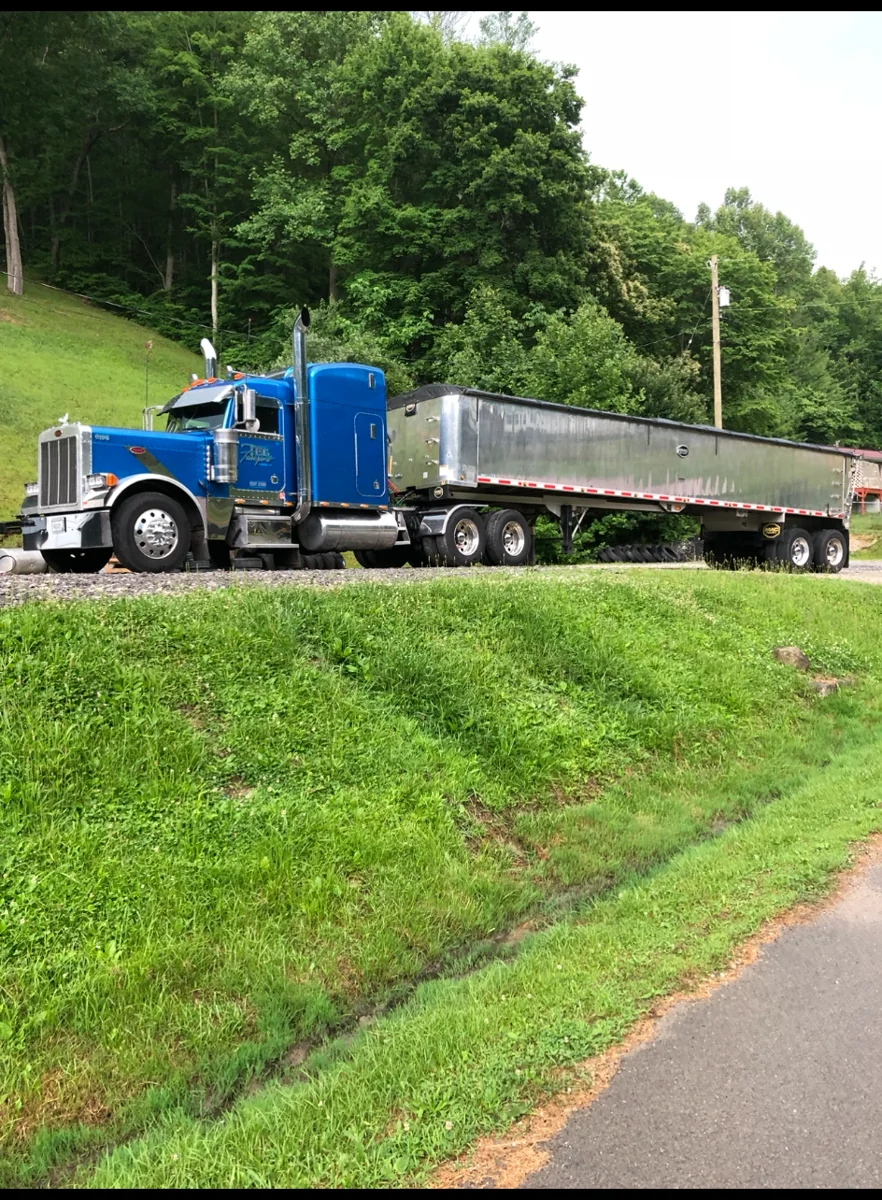 Blue Peterbilt tractor and aluminum dump trailer beside a green hillside